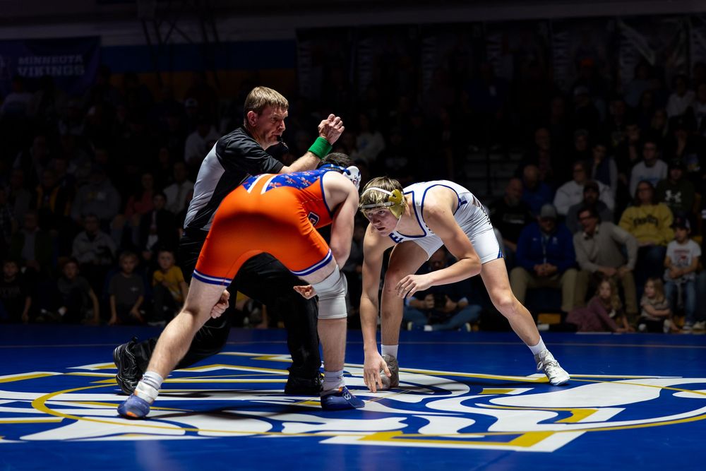 Two wrestlers compete on a blue mat during a wrestling match, watched by a referee and crowd.