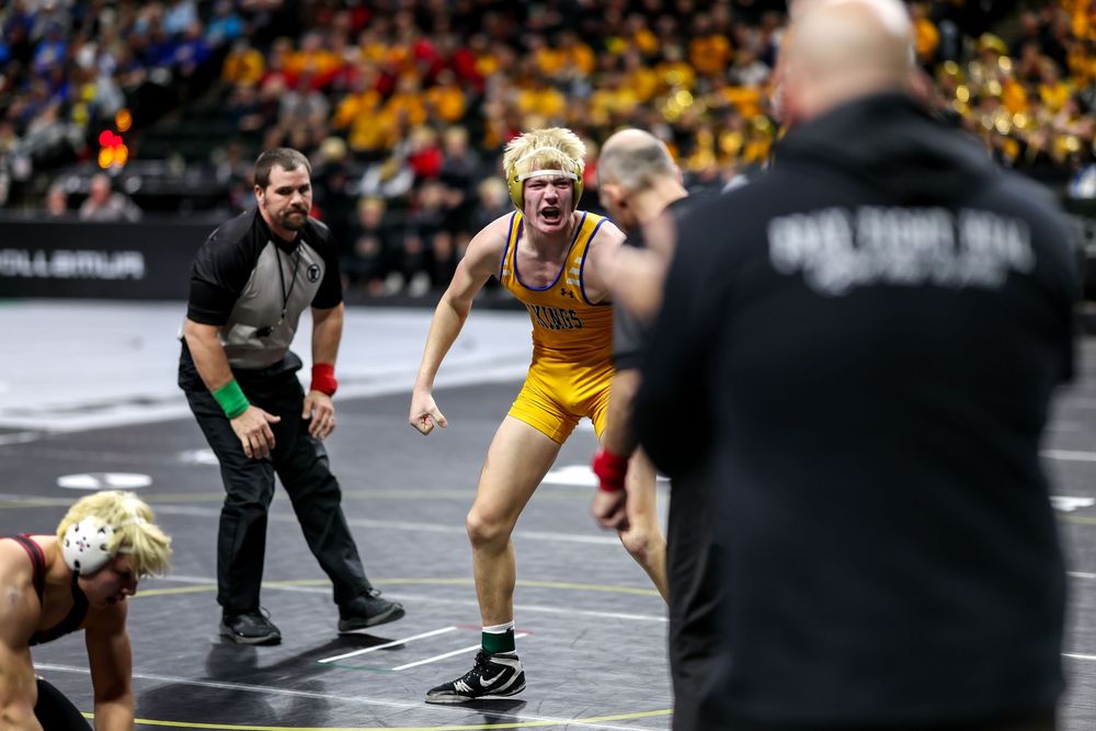 Wrestler in gold singlet celebrates victory on mat, referee and coach visible in arena crowd background.