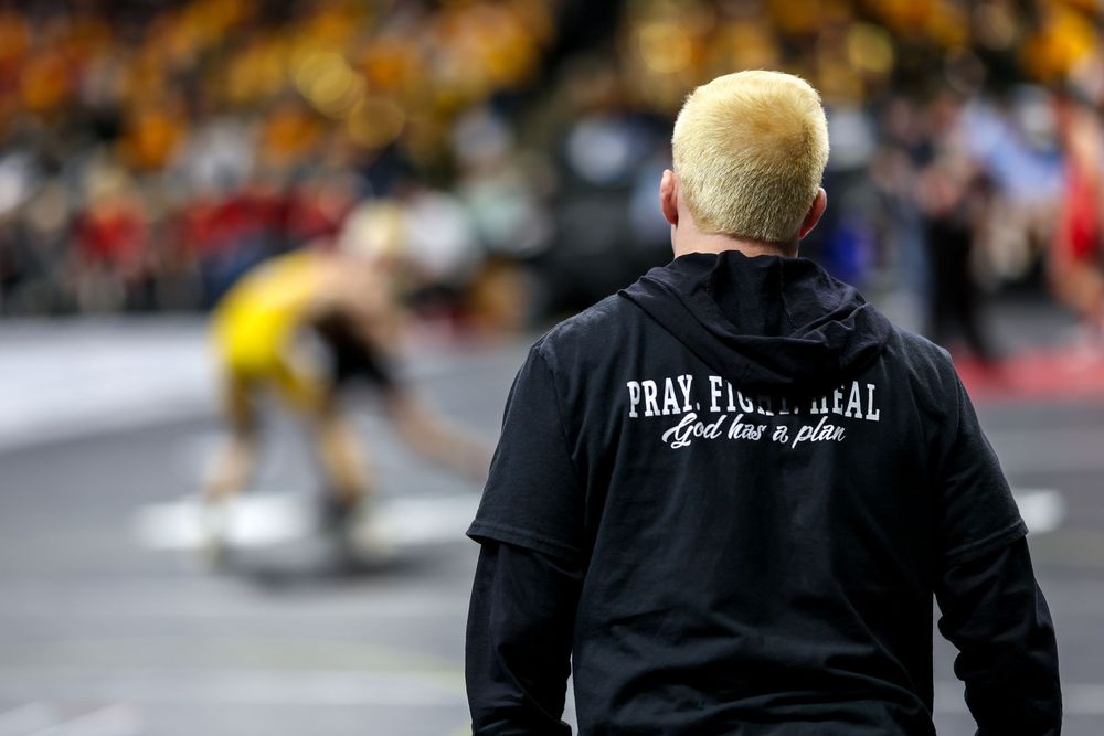 Coach in black hoodie reading Pray Fight Heal watches wrestlers compete on mat at indoor wrestling tournament.