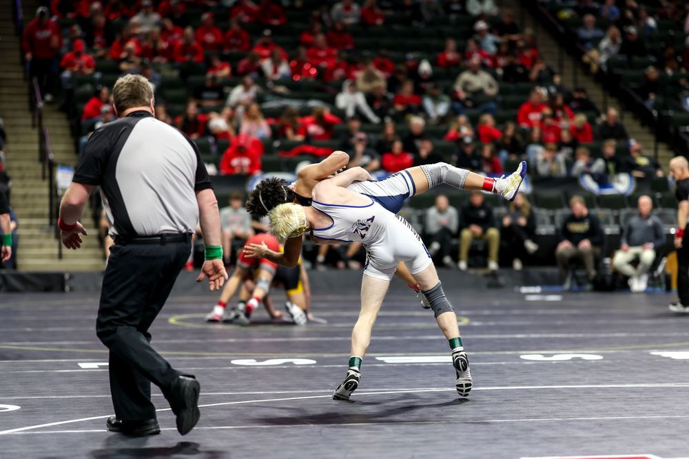 Two wrestlers compete on a mat during a wrestling tournament, with a referee watching closely in an arena.