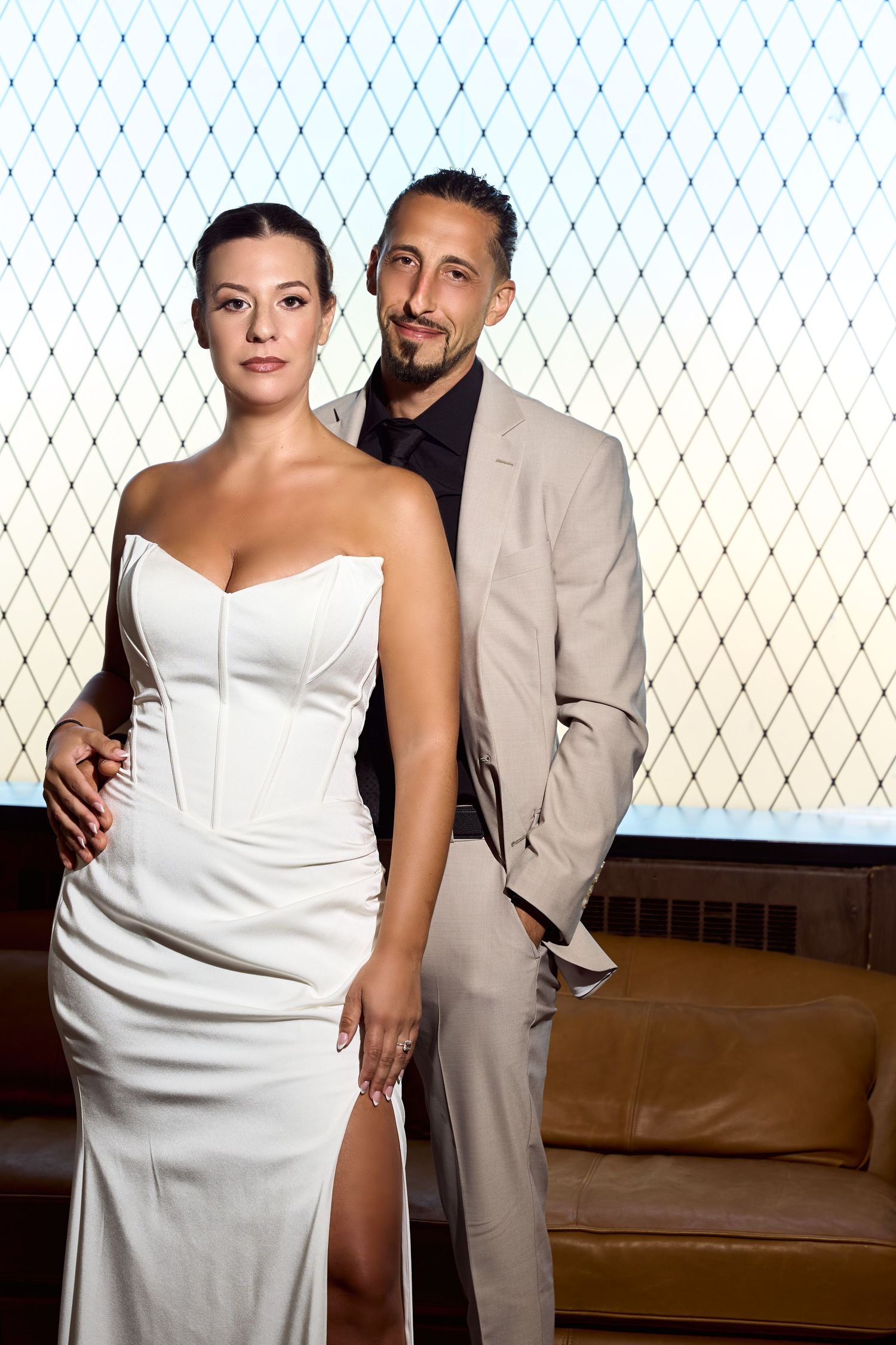 Elegant couple posing formally, woman in white strapless gown, man in beige suit, against geometric window backdrop.