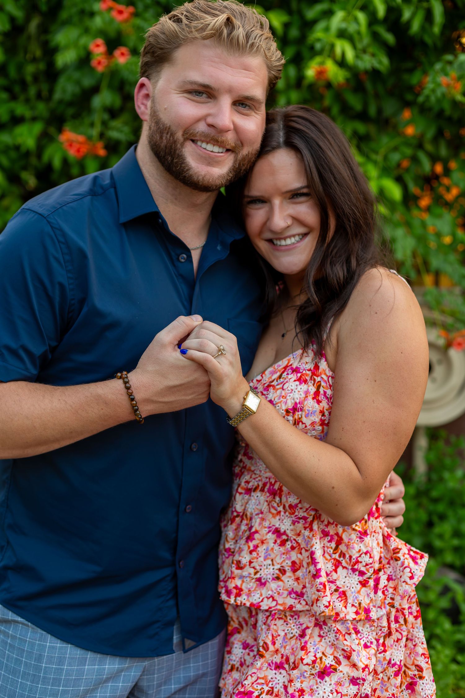 A smiling couple embraces outdoors, the man in a blue shirt and the woman in a floral dress, with colorful flowers behind them.