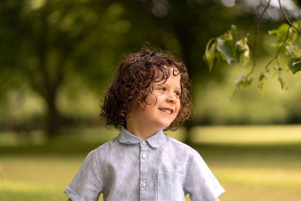 Curly-haired boy in a light blue linen shirt smiling outdoors in a sunny green park.