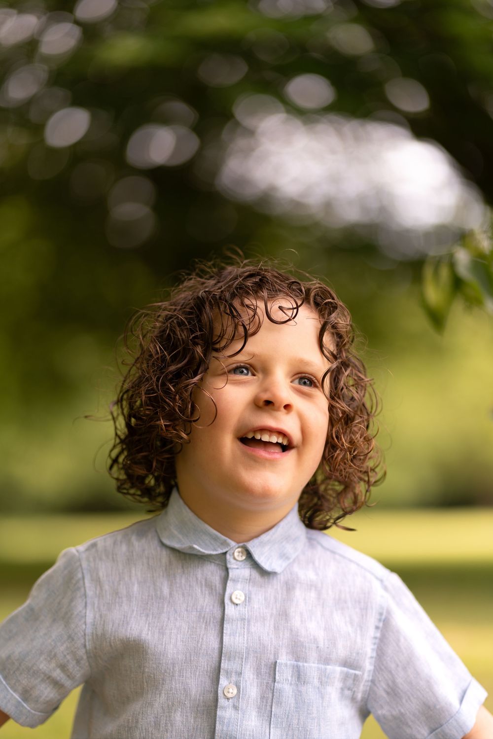 Curly-haired boy in a light blue shirt laughing outdoors in a green park on a sunny day.