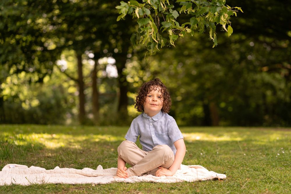 Curly-haired child sitting cross-legged on blanket in sunny green park, looking up thoughtfully.
