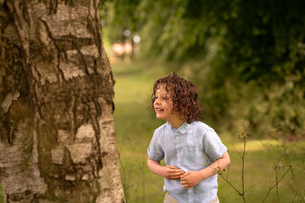 A joyful curly-haired child laughing outdoors near a birch tree in a lush green park setting.