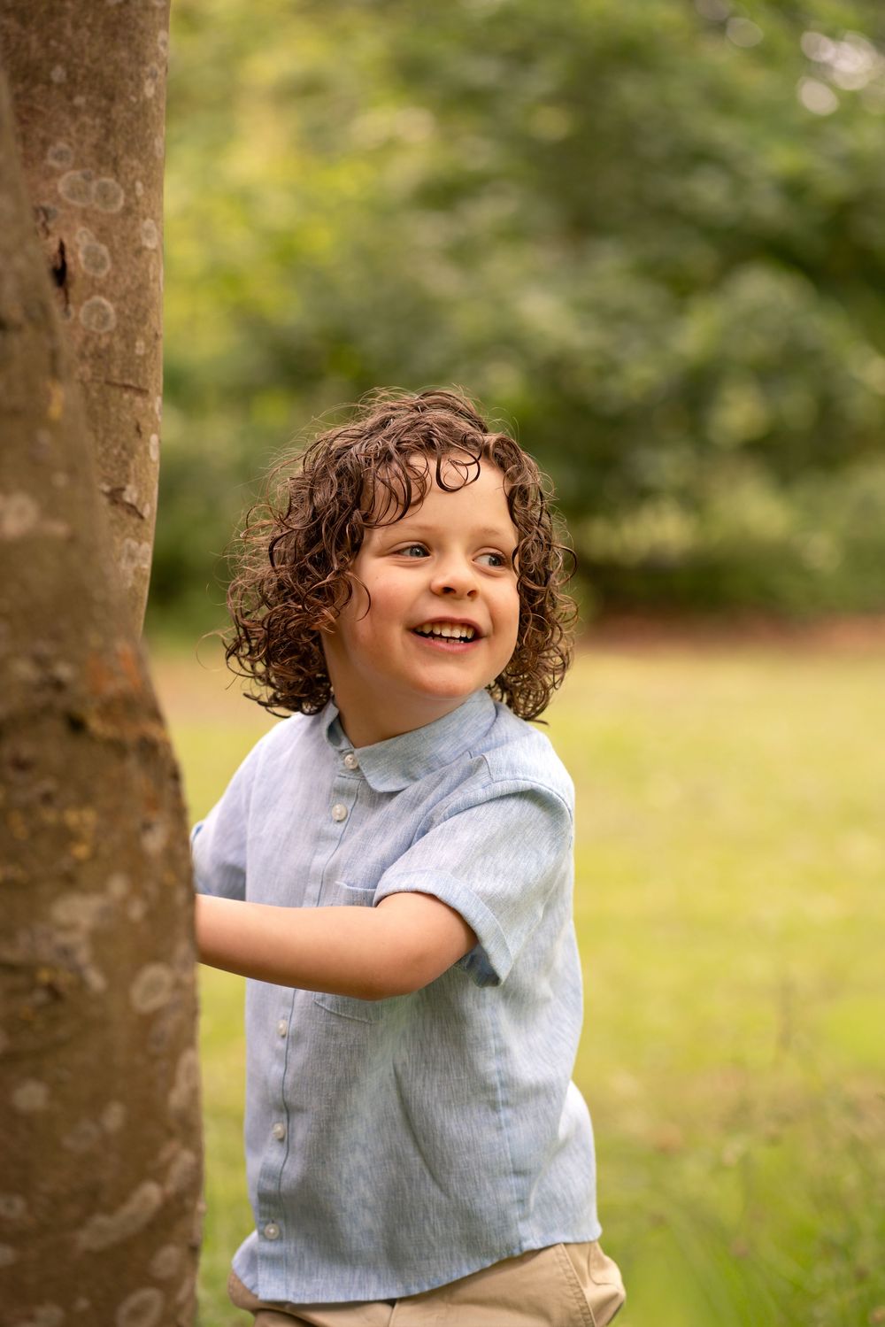 A smiling curly-haired boy in a blue shirt leans against a tree trunk in a lush green park.