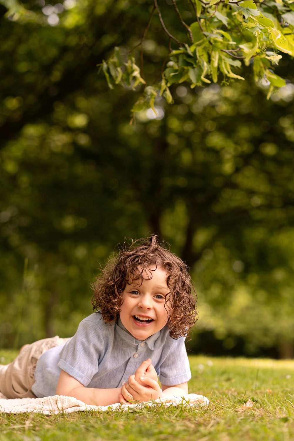 Happy curly-haired boy lying on grass in a sunny park, smiling joyfully while relaxing outdoors.