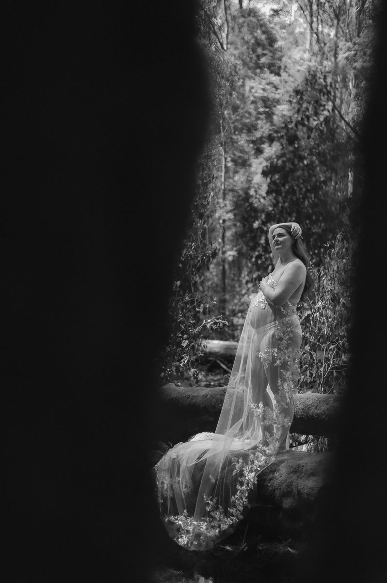 Black and white maternity photo of a pregnant woman in a flowing lace gown standing in a lush forest setting.