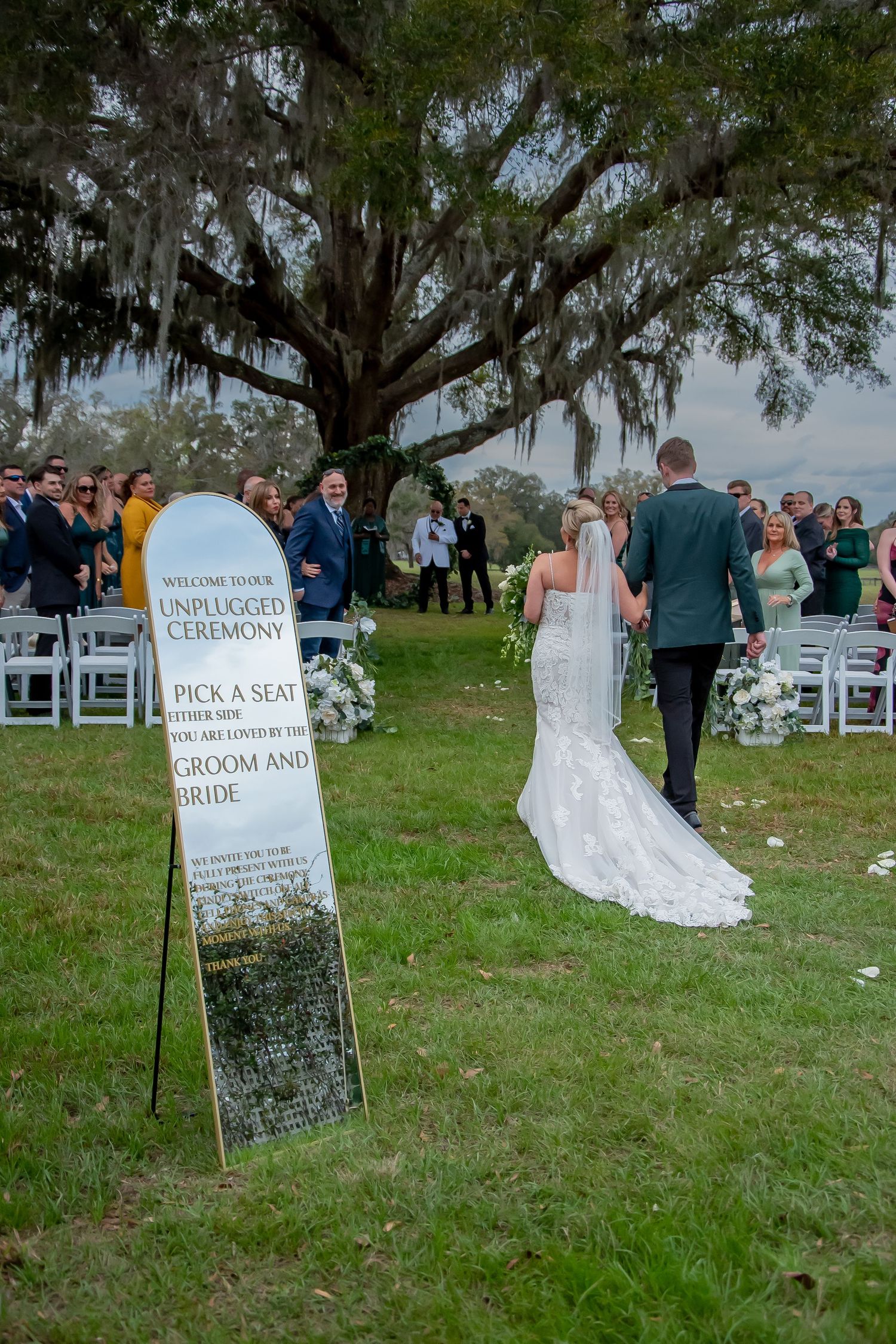 Bride and groom walk down outdoor wedding aisle past unplugged ceremony sign under Spanish moss oak tree.