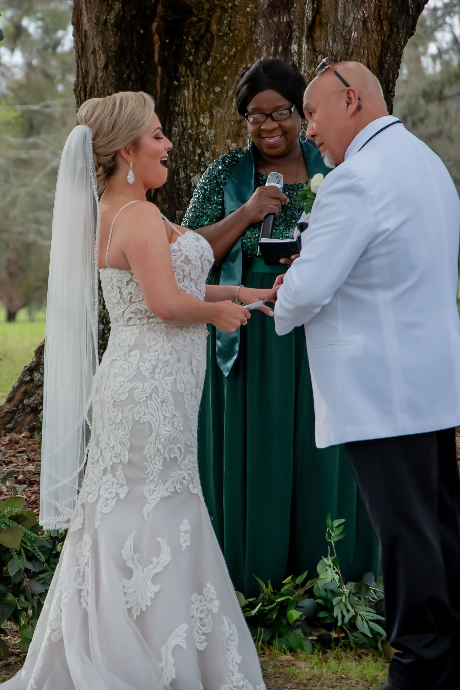 Bride and groom exchange vows with smiling officiant in green gown during outdoor wedding ceremony.