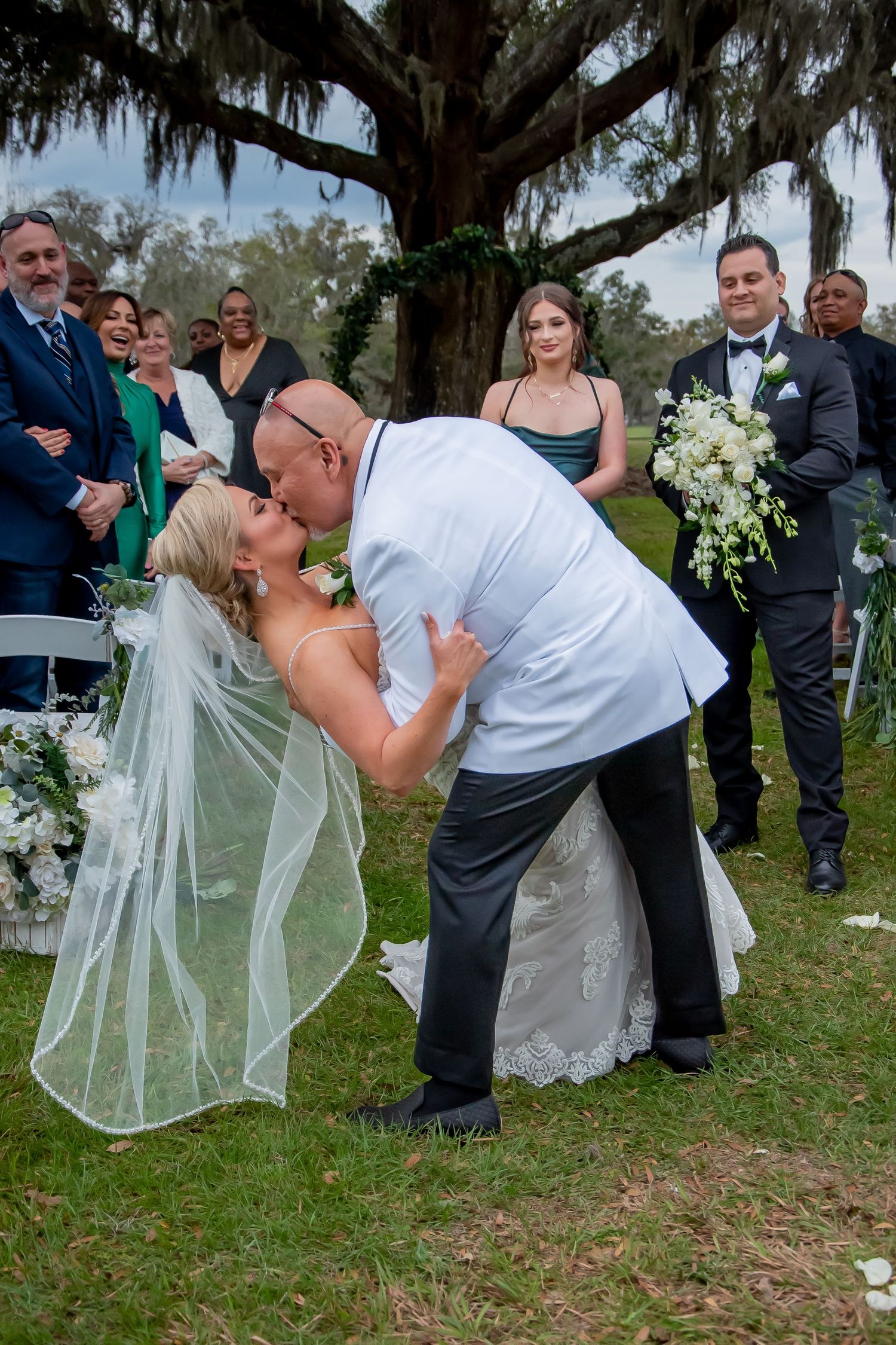 Groom dips bride for romantic kiss at outdoor wedding ceremony under oak tree with guests watching.