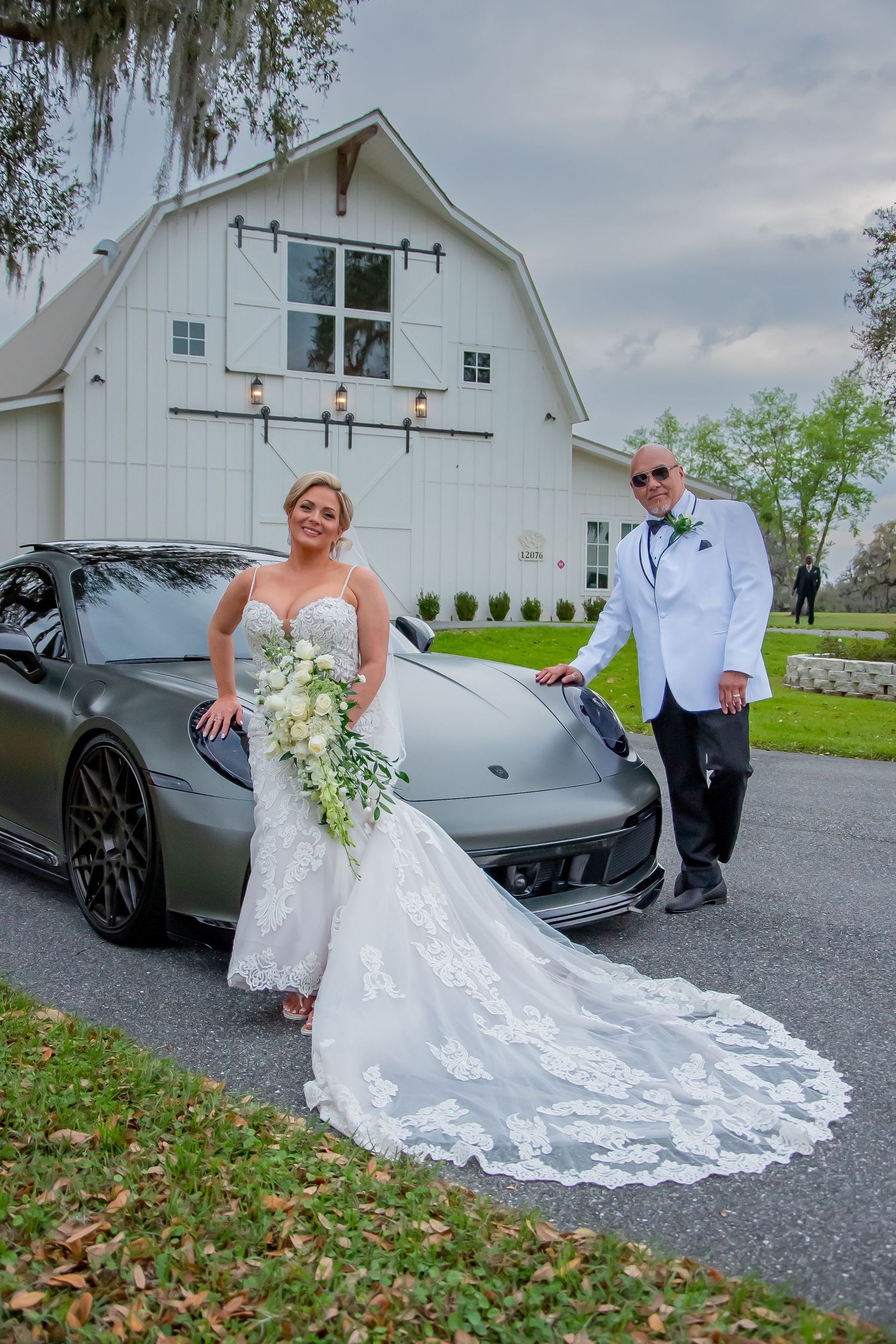 Bride and groom pose with gray Porsche sports car in front of white barn wedding venue.