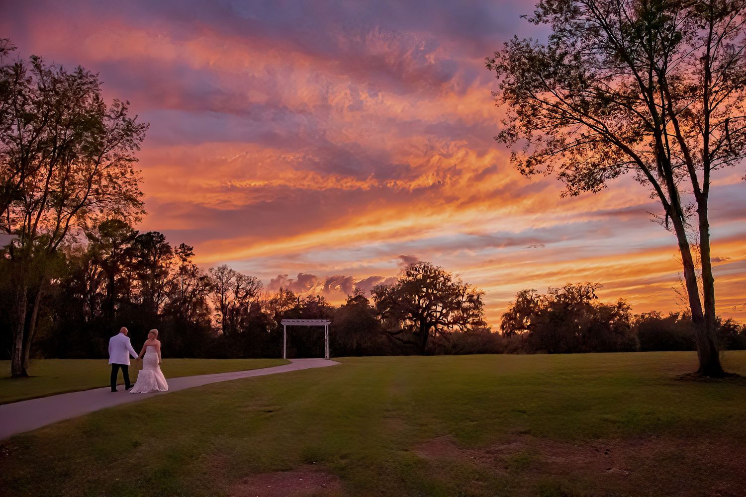 Couple walks toward garden gazebo under a stunning fiery sunset sky with vibrant orange and purple clouds at outdoor wedding.