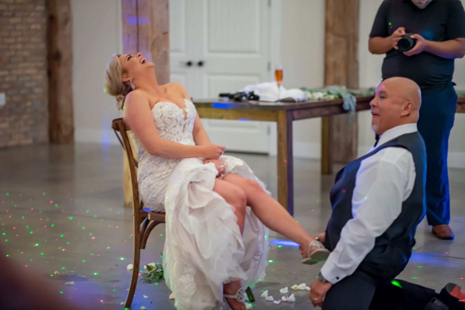 Groom removes bride's garter during fun wedding reception tradition on dance floor with colorful lights.