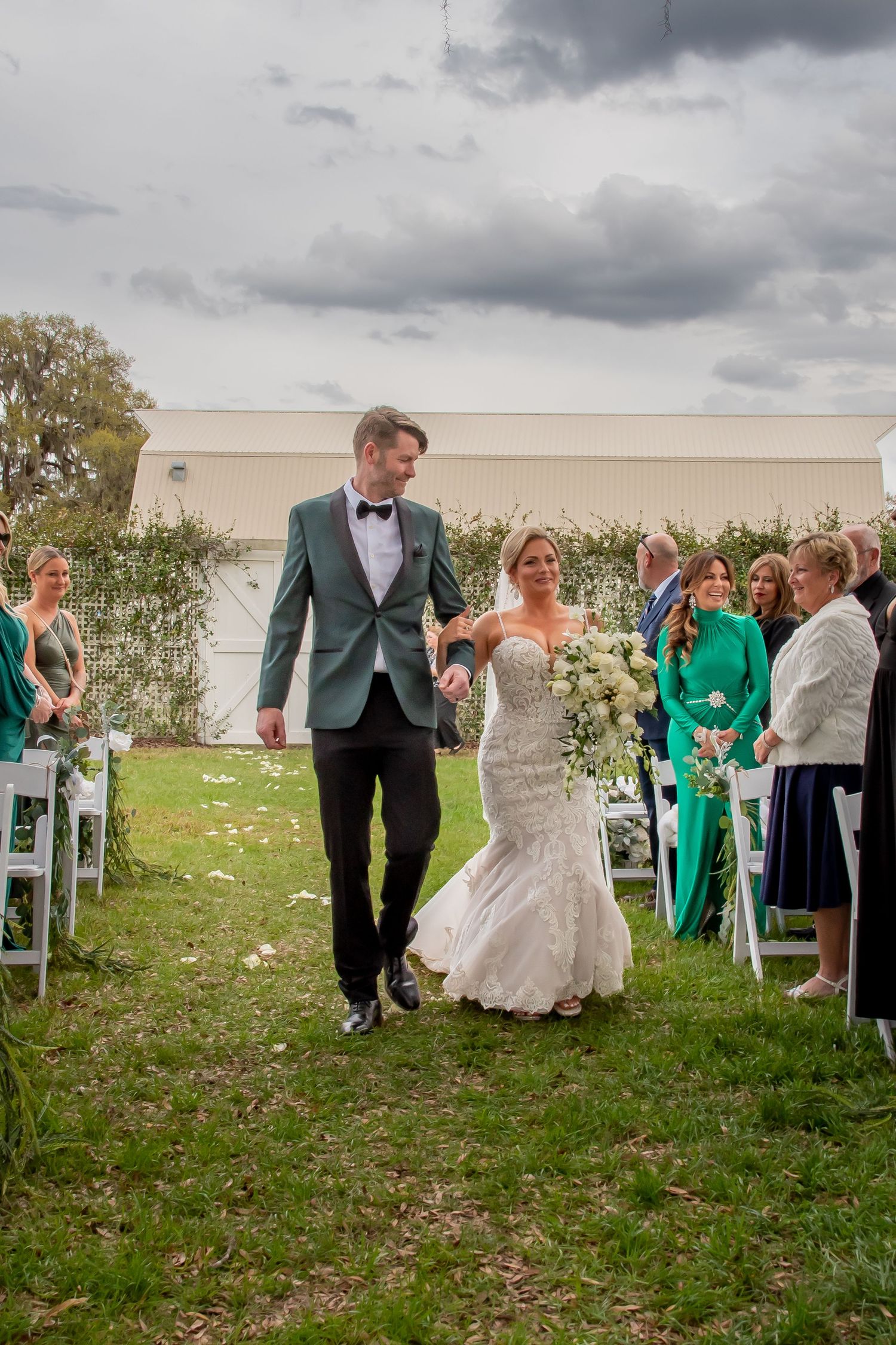 Bride and groom walk down outdoor wedding aisle holding hands, bride in lace mermaid gown holding white bouquet.