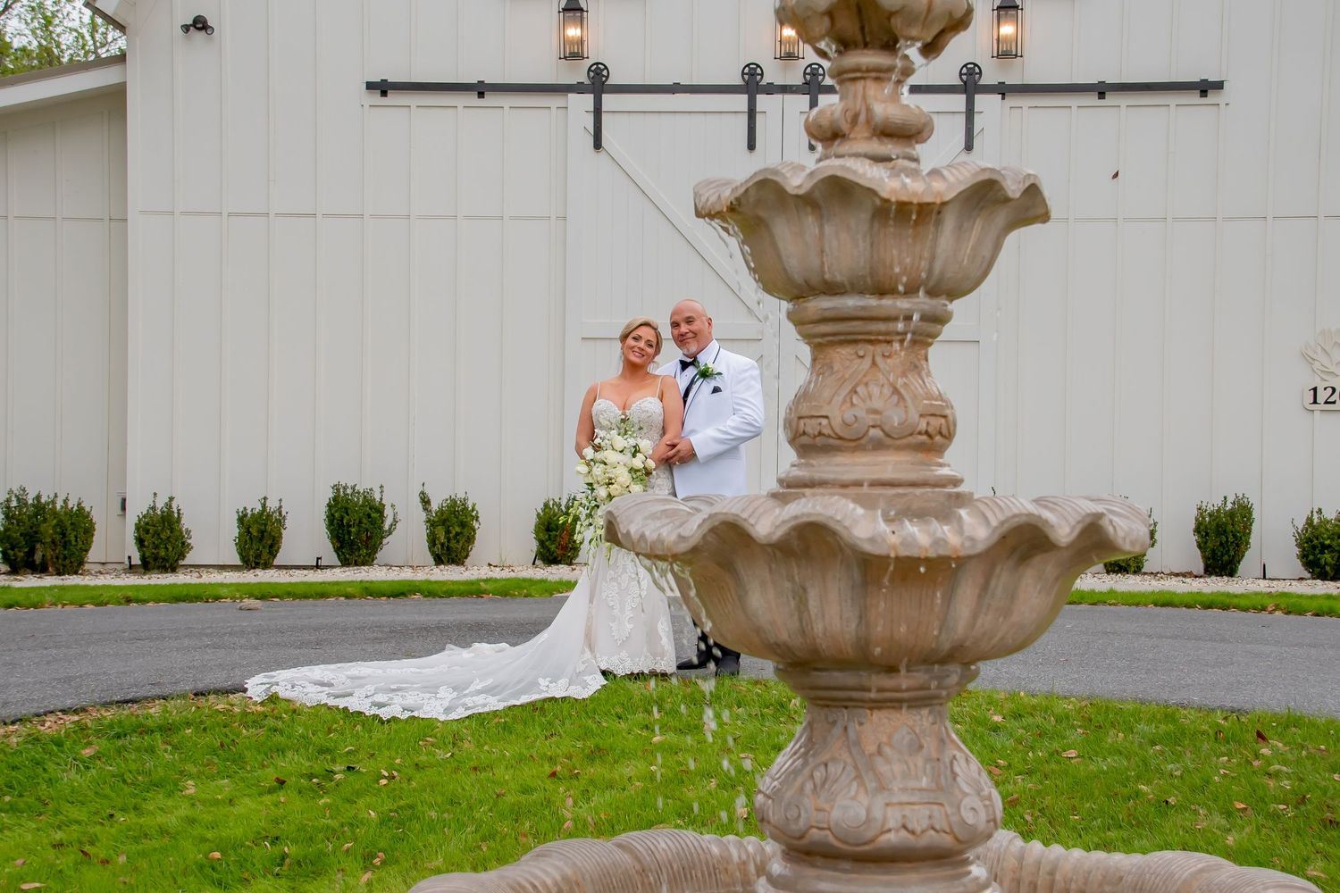 Bride and groom pose romantically behind an ornate stone fountain outside a white barn wedding venue.