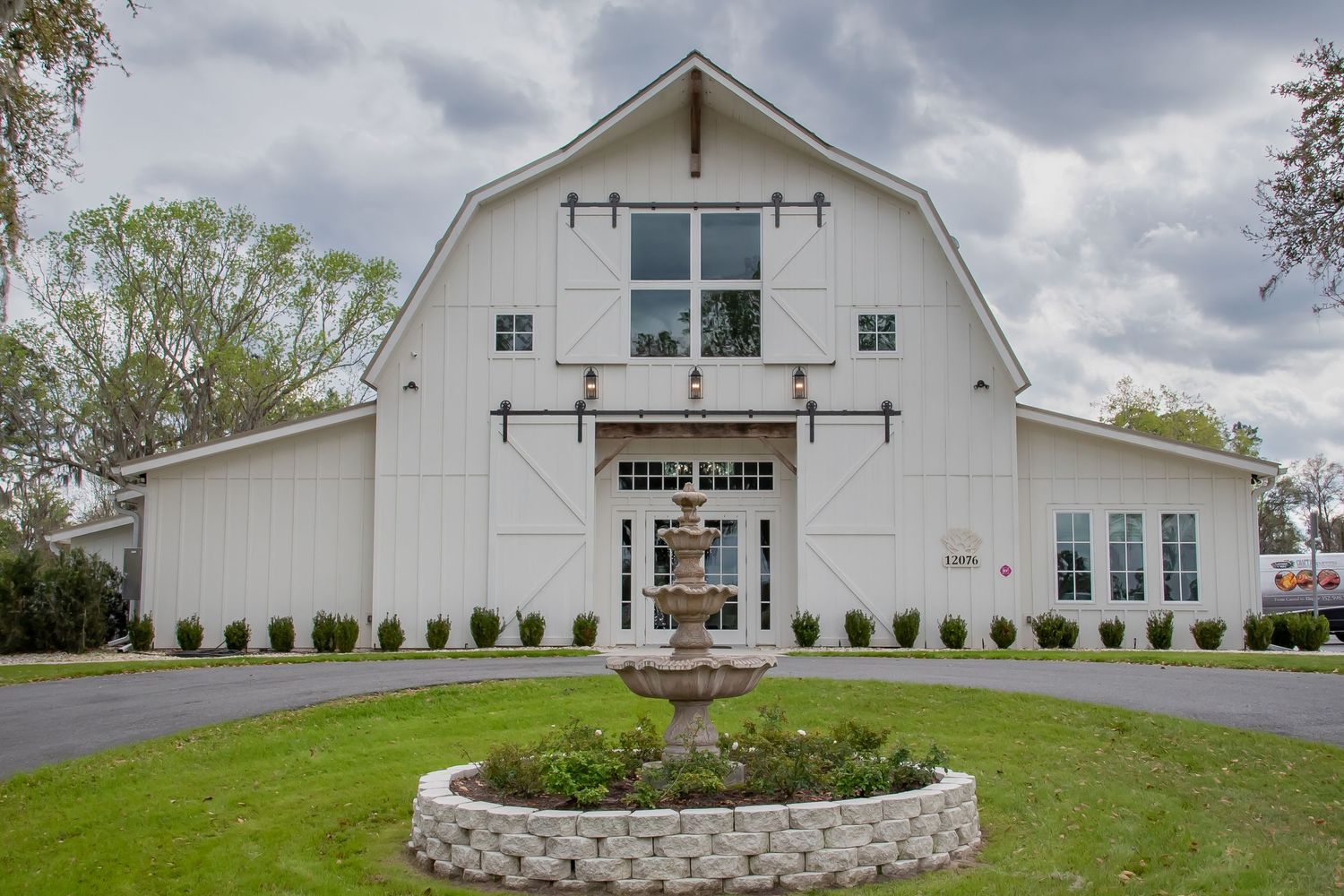 White barn-style wedding venue with sliding doors, circular garden fountain, and lush green lawn under cloudy skies.