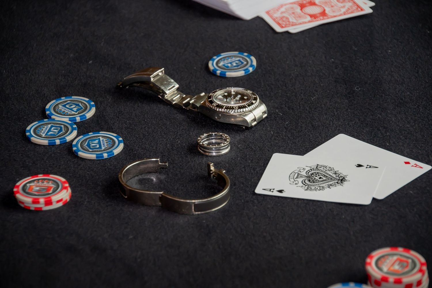 Poker table with casino chips, a luxury watch, bracelet, and playing cards including an ace on dark felt.
