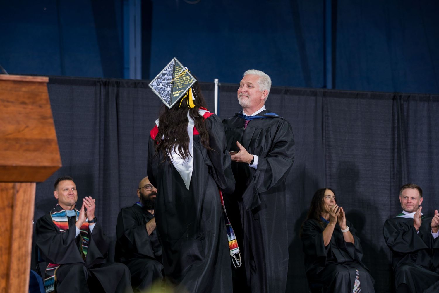 Graduate receives diploma on stage at commencement ceremony, shaking hands with official in academic regalia.
