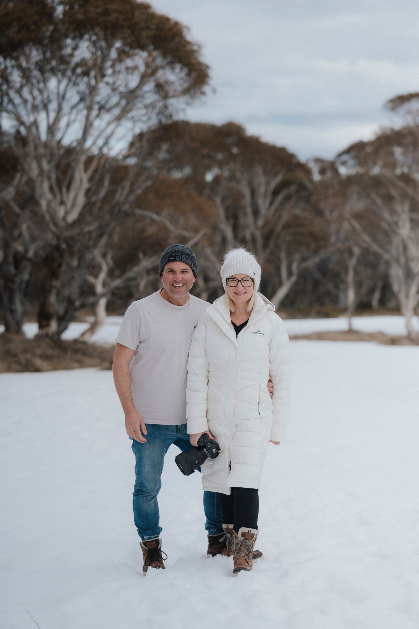 Couple smiling in snowy landscape, woman in white puffer jacket, man holding camera.