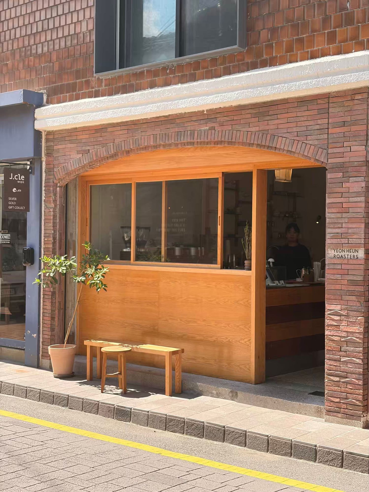Cozy wooden cafe storefront with arched brick exterior, large windows, and small stools on a sunny sidewalk.