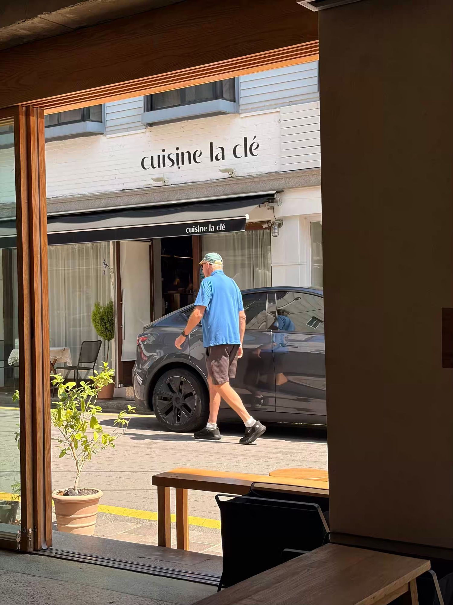 Man in blue shirt walking past Cuisine La Clé restaurant with a black car parked outside on a sunny day.