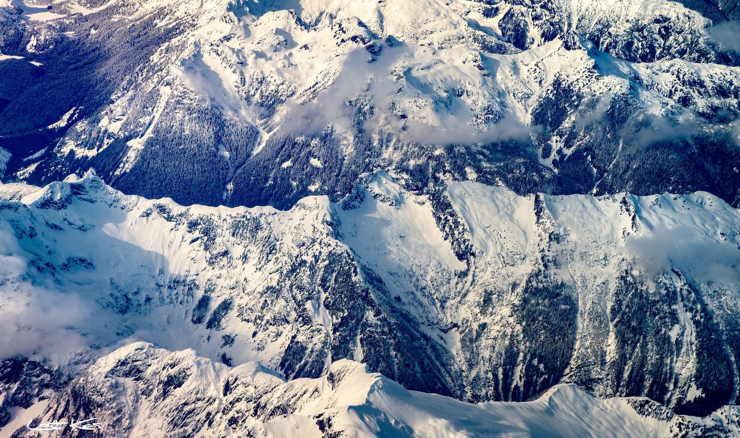 Snow-covered mountain peaks of a vast mountain range viewed from high above through scattered clouds.