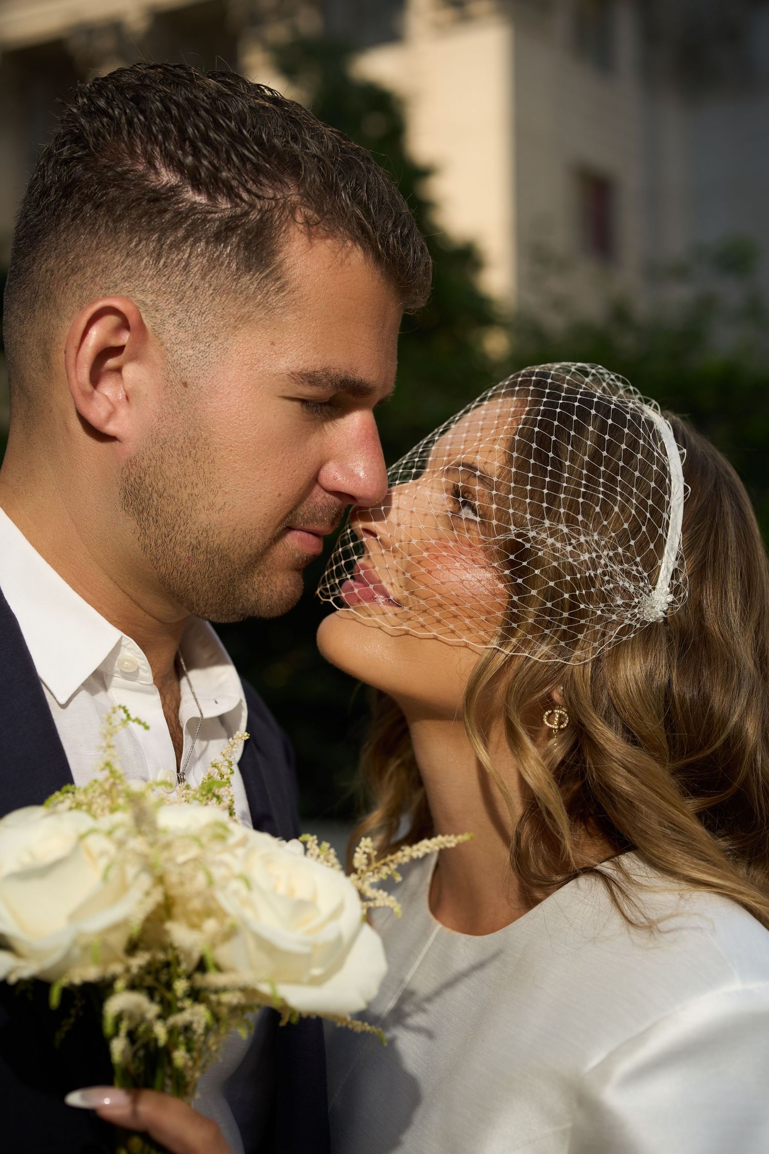Bride in birdcage veil and groom sharing a romantic kiss outdoors, bride holding white rose bouquet on wedding day.