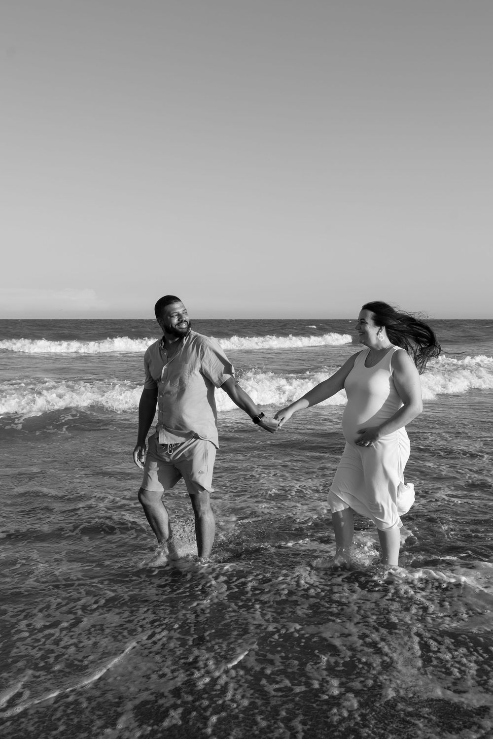 Black and white photo of a couple holding hands and walking together in ocean waves on a sunny beach.