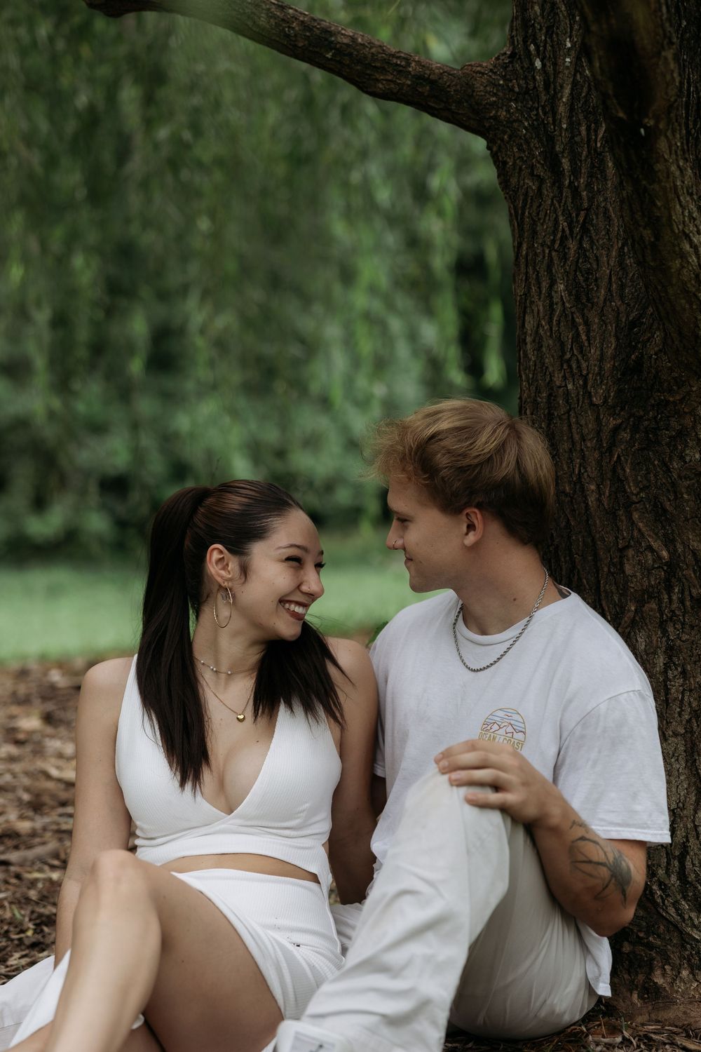 A couple sits together under a tree in a lush park, smiling at each other, dressed in matching white outfits.