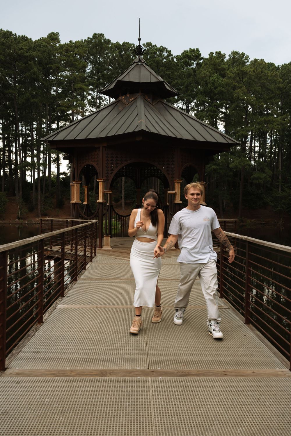 Couple holding hands walking across a bridge toward a gazebo surrounded by tall pine trees.