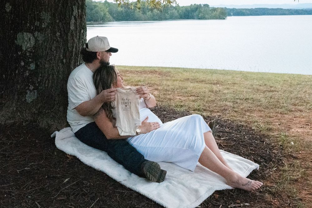 Couple sitting against a tree by a lake, holding a baby onesie, enjoying a peaceful outdoor maternity moment.