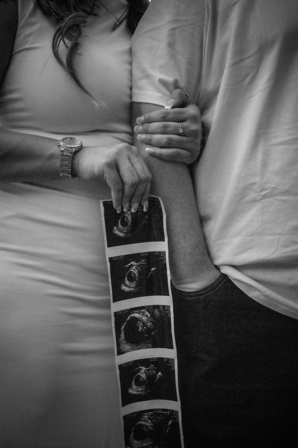 Black and white maternity photo of couple holding ultrasound strip, woman in fitted dress with watch, man in white shirt.