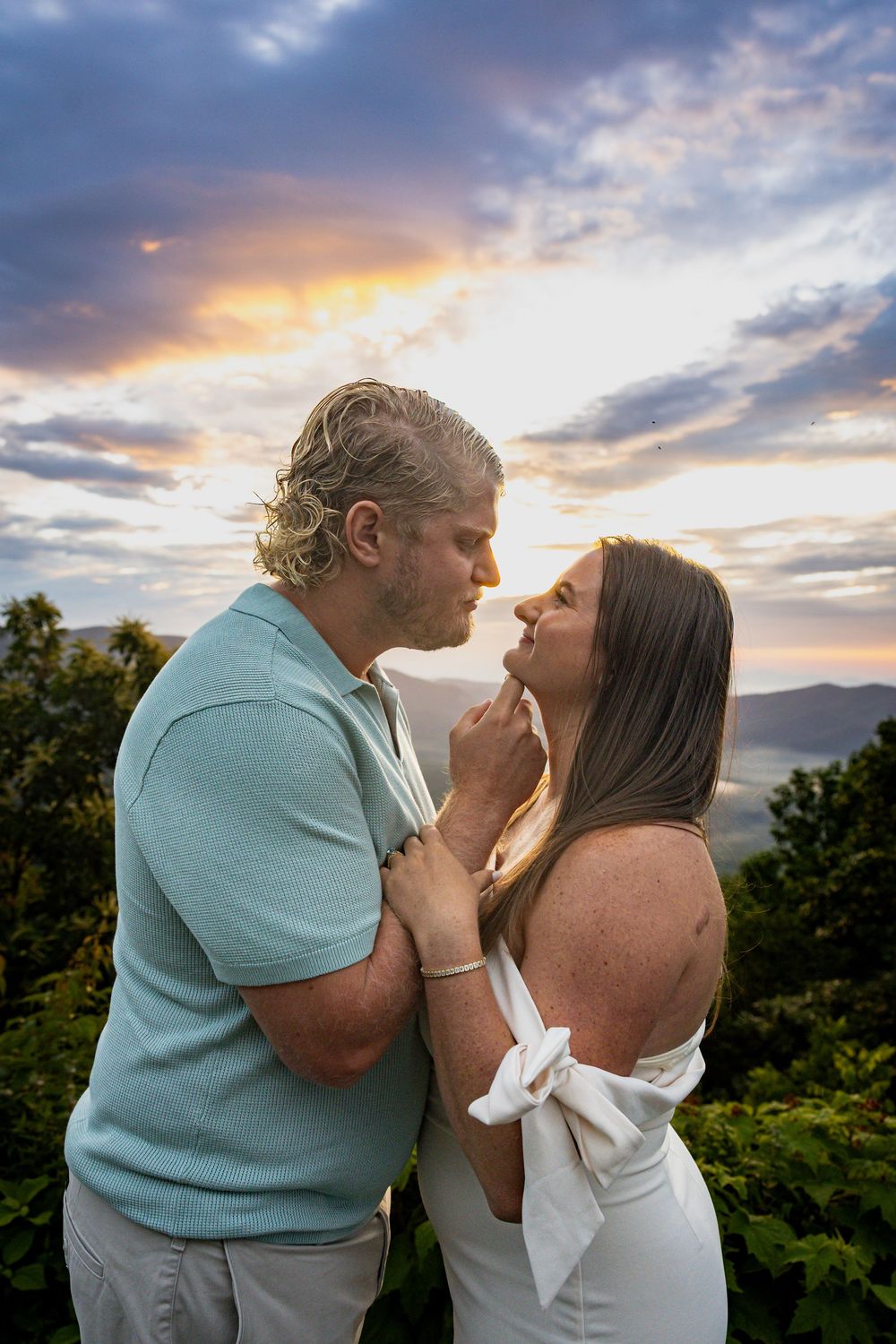 Couple sharing a romantic moment at sunset with dramatic clouds and mountain views in the background.