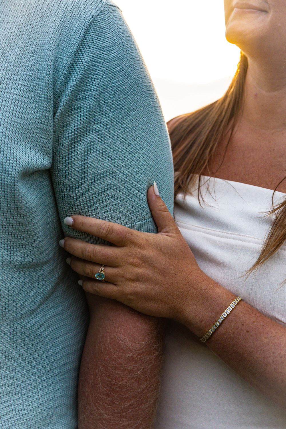 Woman with engagement ring and gold bracelet holding man's arm in teal shirt at sunset.