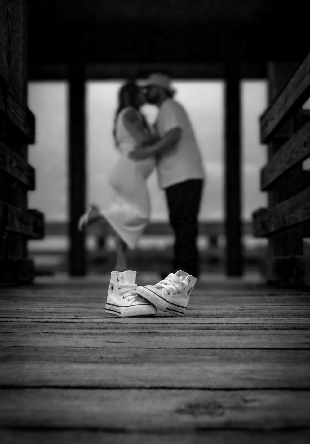 Black and white maternity photo of couple kissing on pier with tiny Converse sneakers in foreground.