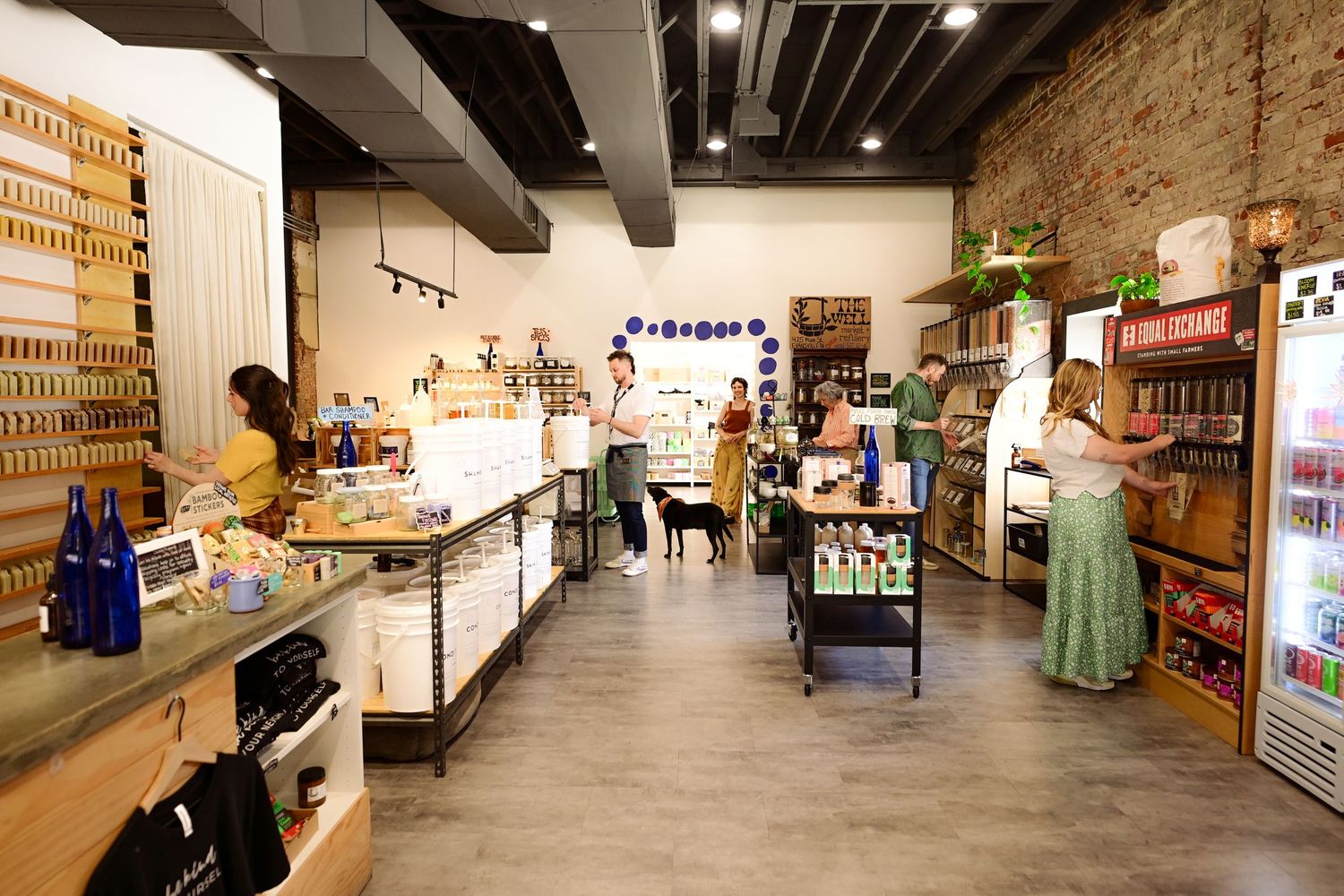 Bright retail store interior with shoppers browsing shelves of products, exposed brick wall, and wood floors.