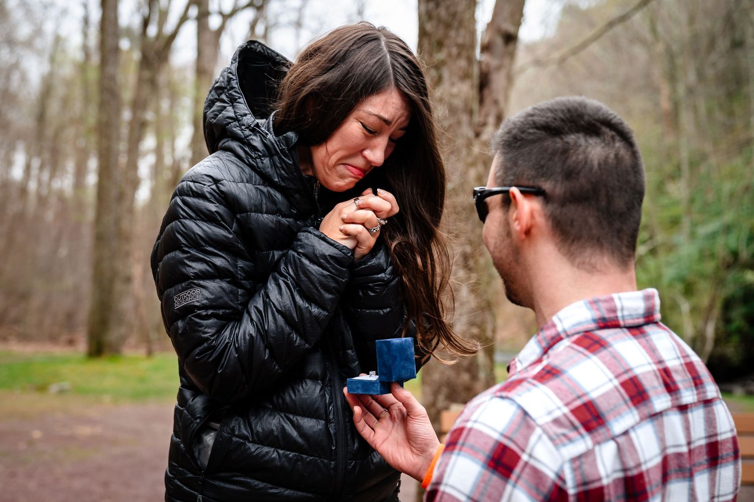 Man kneeling on one knee proposing to emotional woman with ring box outdoors in wooded park setting.