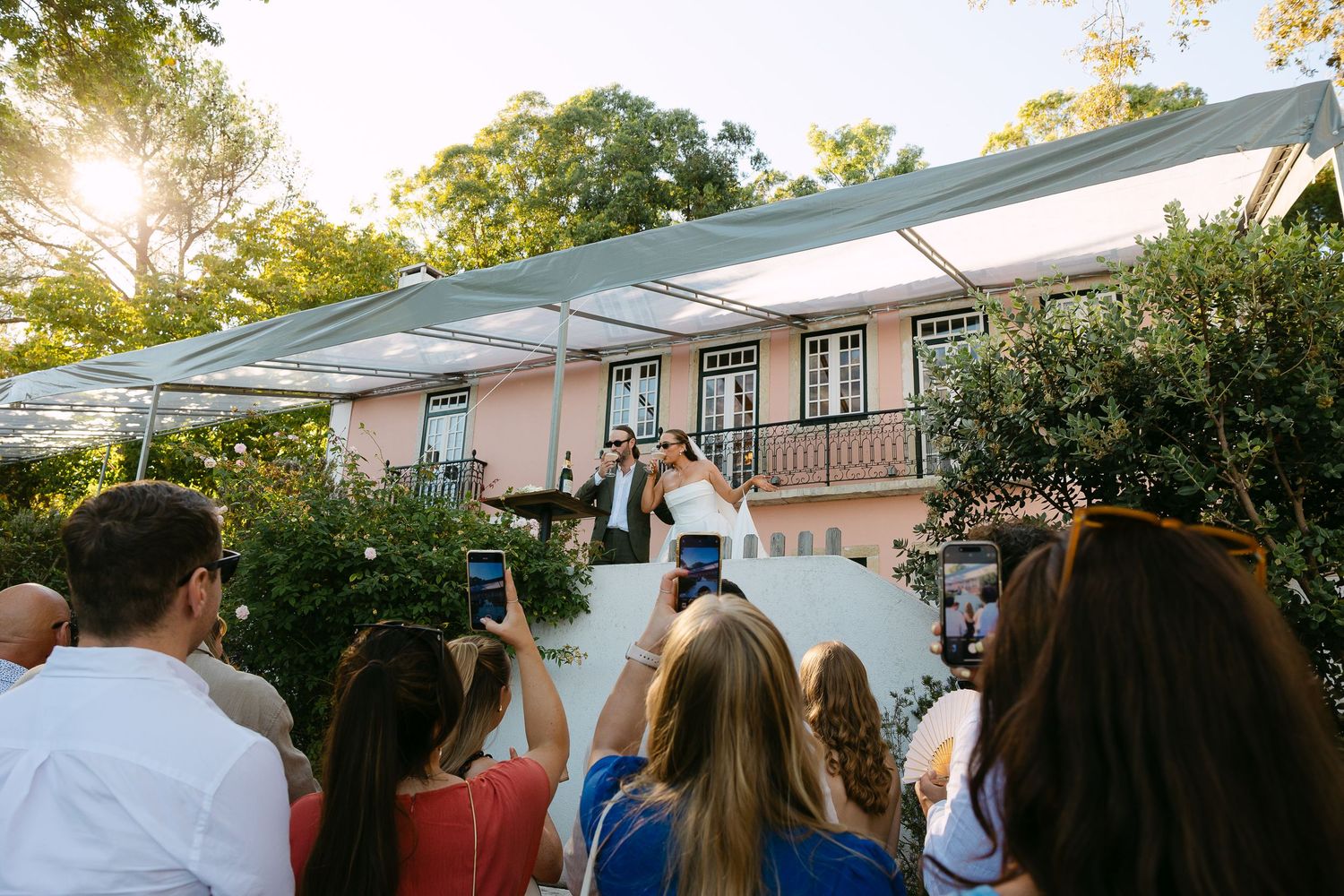 Couple kisses on steps of pink venue while guests photograph them outdoors at a sunny wedding celebration.