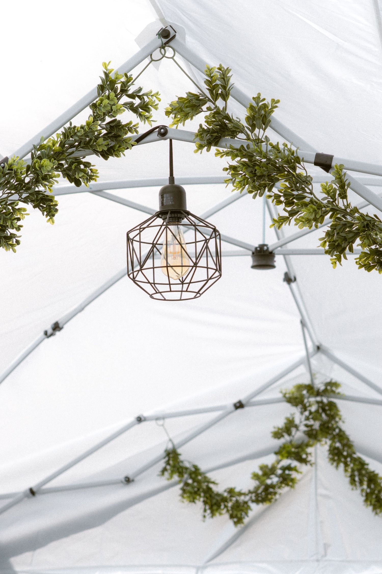Geometric cage pendant light hanging from white tent frame decorated with green leaf garlands.