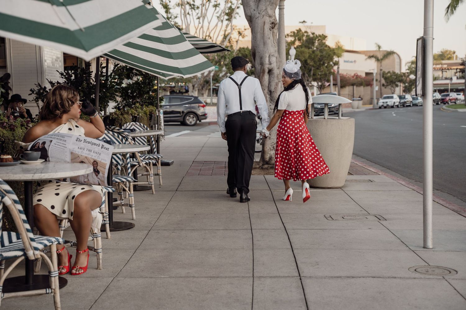 A couple in retro 1950s attire walk past an outdoor cafe with striped awnings on a sunny street corner.