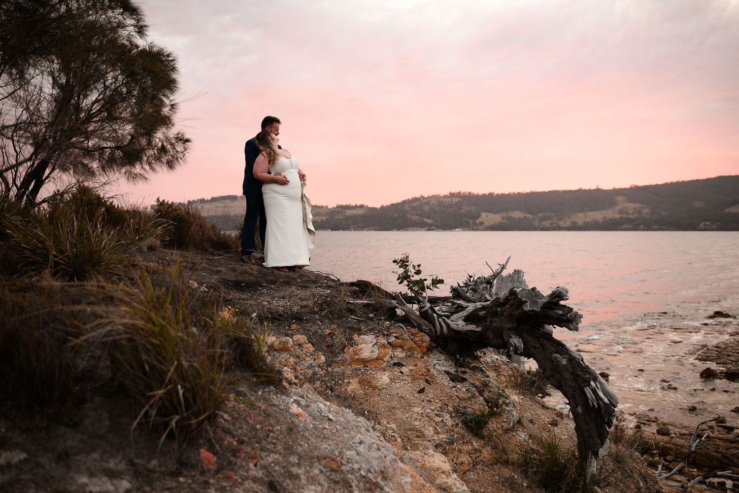 A couple stands together on a rocky coastal cliff overlooking a serene body of water at sunset.