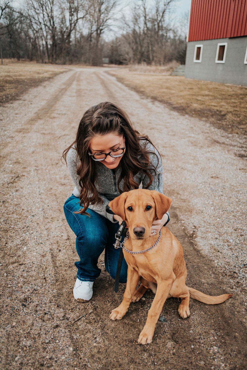 Person in blue jacket kneeling to pet brown dog on gravel path.