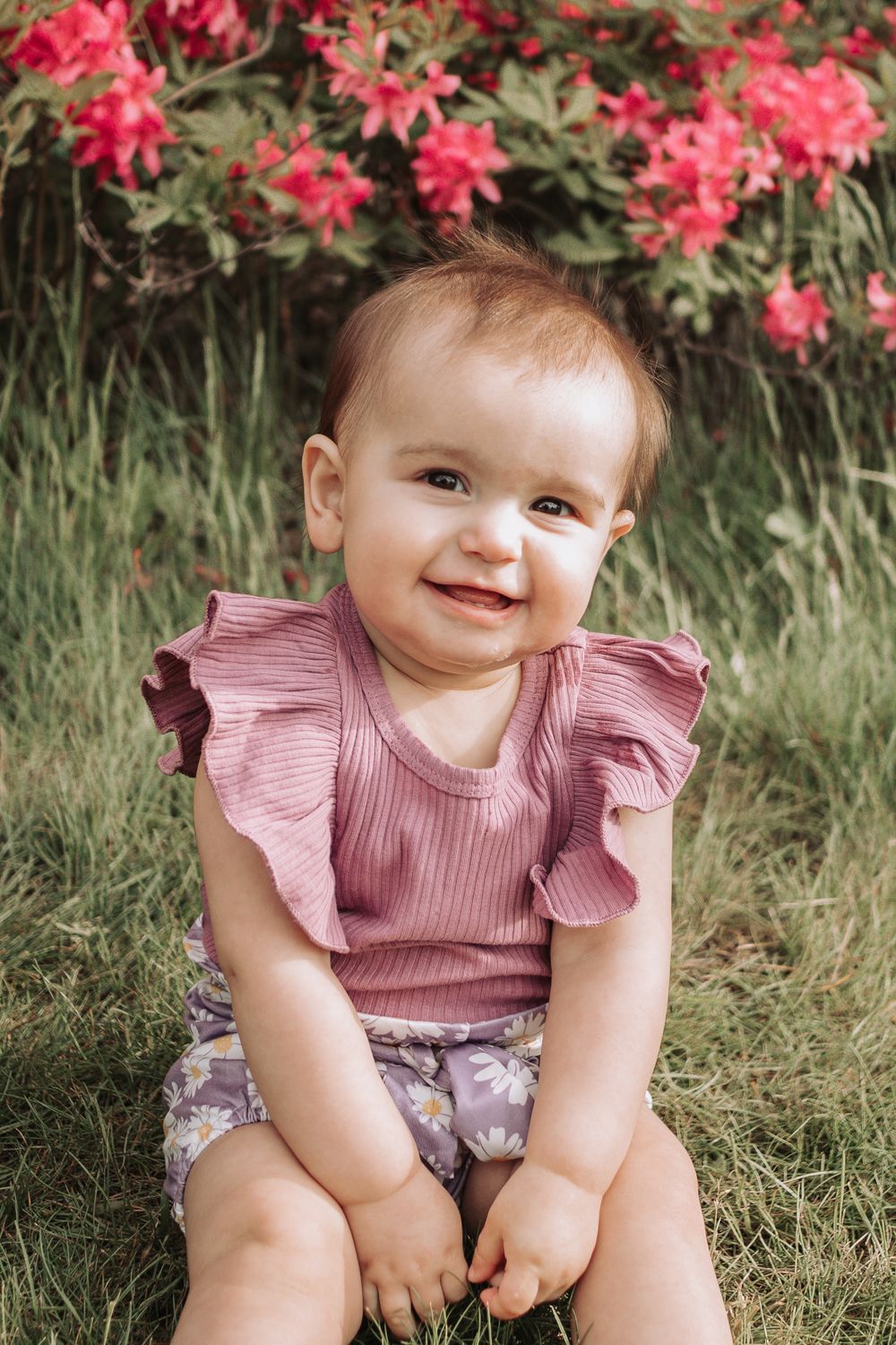 Baby in pink ruffled top smiling while sitting in front of vibrant pink flowers.