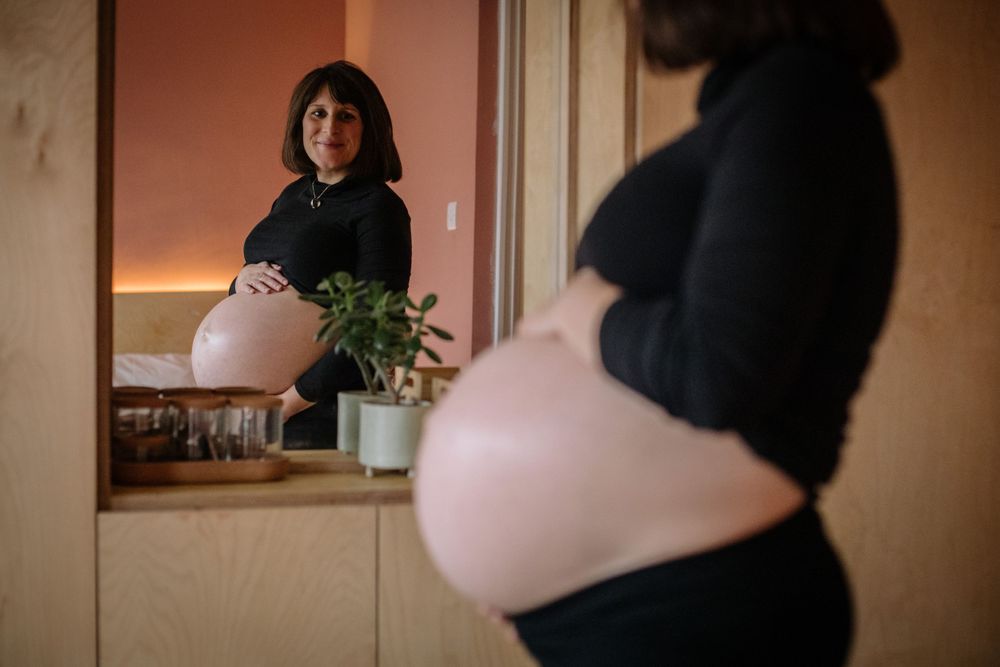 Pregnant person wearing dark clothing looks into mirror while standing in warmly lit room with houseplant.