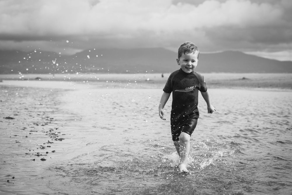 A child splashes through shallow water at the beach in a black and white photo with mountains in the distance.