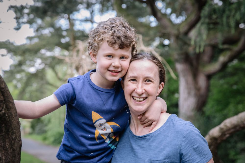 A child with curly hair wearing a blue t-shirt hugs their parent while smiling together outdoors near trees.