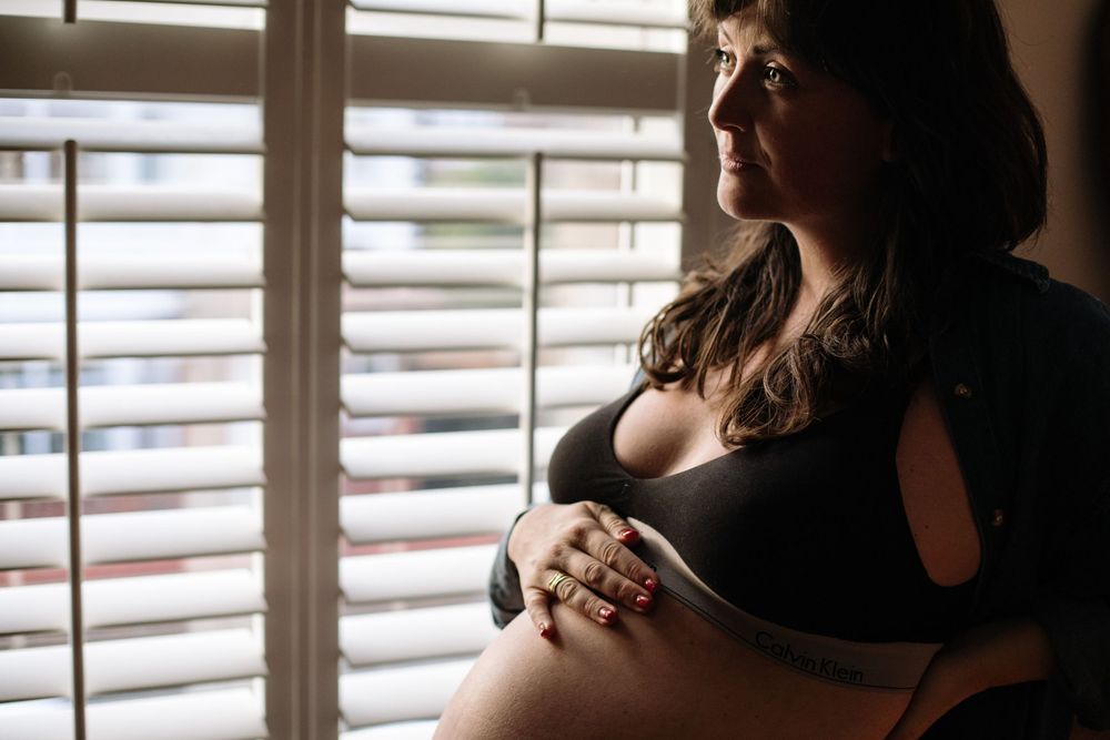 A silhouetted pregnant woman in a black top poses by a window with venetian blinds casting dramatic light.