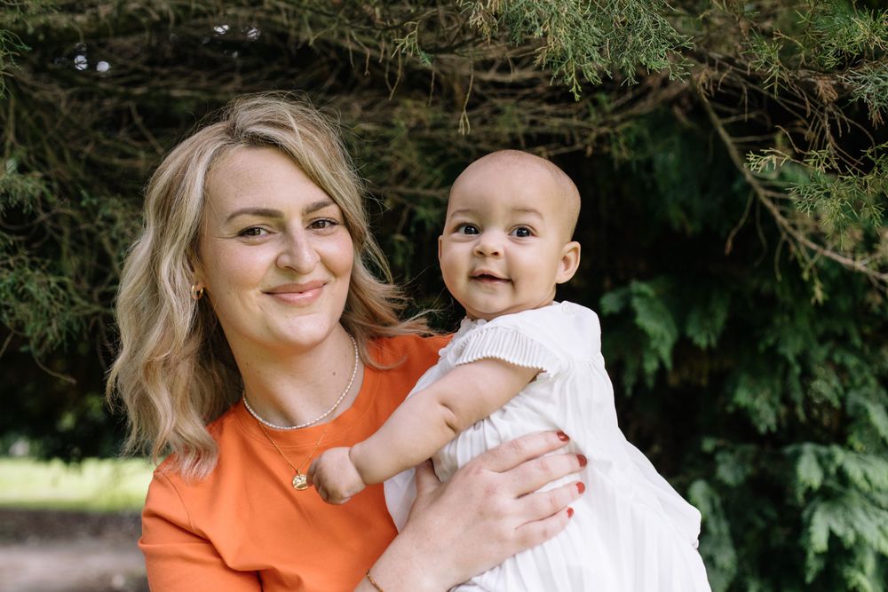 A smiling person in orange shirt holds a baby in white outfit against a backdrop of green foliage outdoors.
